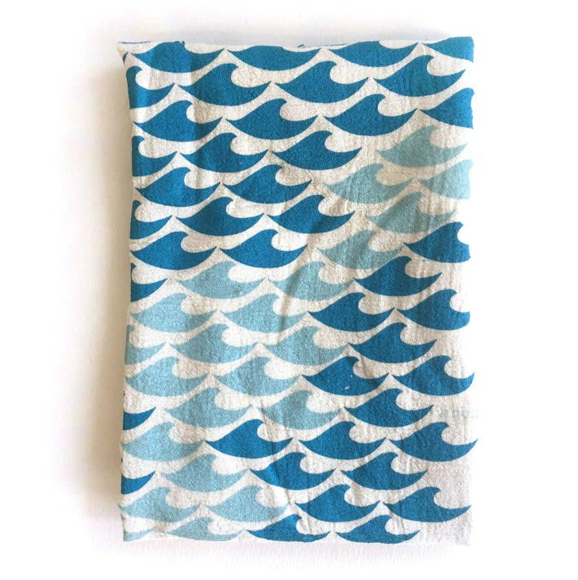 A folded beige cotton tea towel with a silkscreened blue and green wave pattern design, placed on a wooden surface.