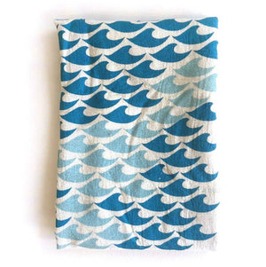 A folded beige cotton tea towel with a silkscreened blue and green wave pattern design, placed on a wooden surface.