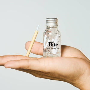 A hand holding a bottle of Bite whitening gel and a bamboo brush. 