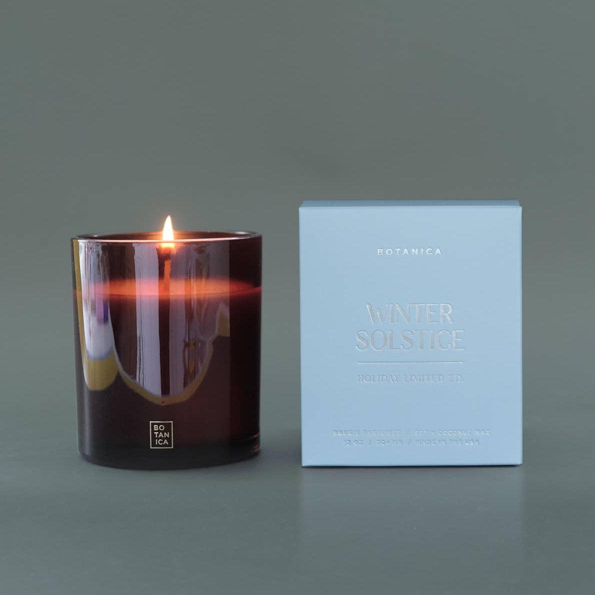 Lit candle in a dark container next to its packaging on a gray background