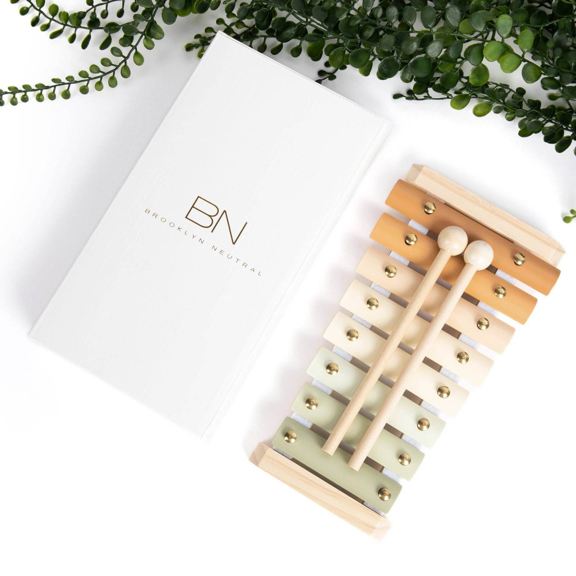 A neutral colored xylophone with wooden keys and metal accents, alongside its packaging box, placed on a white surface with green foliage in the background.